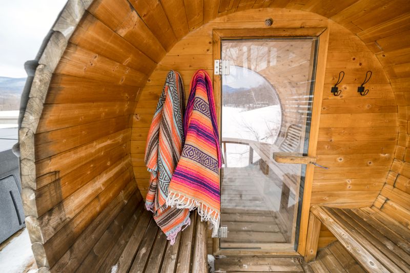 Inside a barrel sauna: two striped woven blankets hanging beside a glass door that opens onto snow, a bench on either side