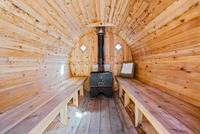 Inside a barrel sauna: pale cedar staves, two facing benches, a black wood-burning stove with its flue rising through the roof, and a basket of folded towels on the rear bench