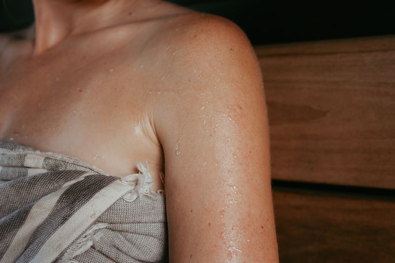 A close-up of a woman's shoulder and upper arm beaded with sweat, a striped linen towel wrapped beneath it, a timber wall behind