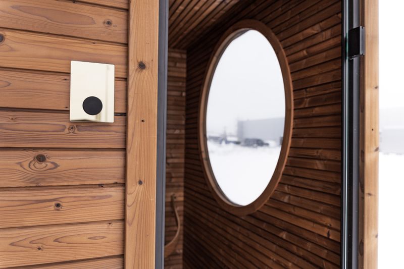 The doorway of a thermo-wood sauna cabin with a round porthole window and a blank control panel on the outer wall, snow outside