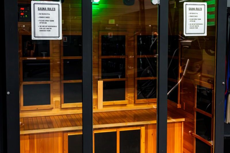 A glass-fronted sauna cabin in warm red-toned timber, flat black heater panels set into the wall above and below the bench, a red digital readout showing 140 at the top left and a printed sauna-rules notice on two of the glass panels